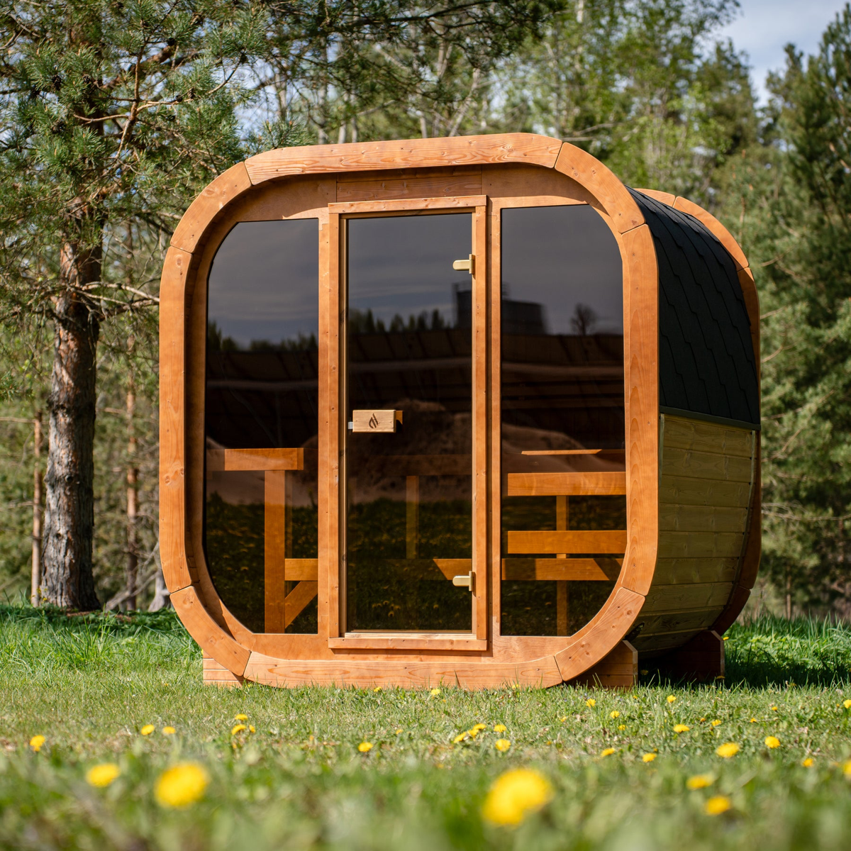 PlusLife Max Traditional Sauna 2.0: rounded-square timber outdoor cabin with a full tinted-glass front, a centred glass door and a black shingle roof, standing on grass in front of pine trees